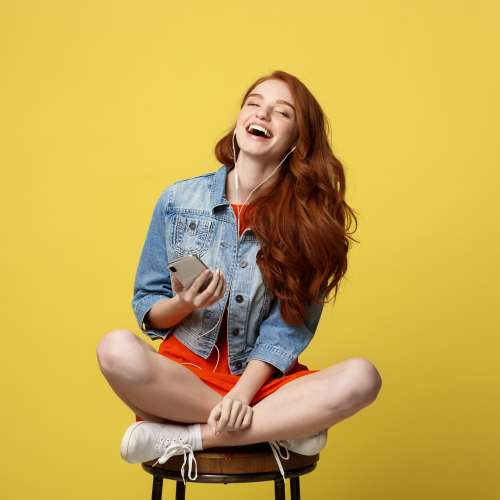 Happy woman in earphones listening to music and singing while sitting and holding mobile phone.